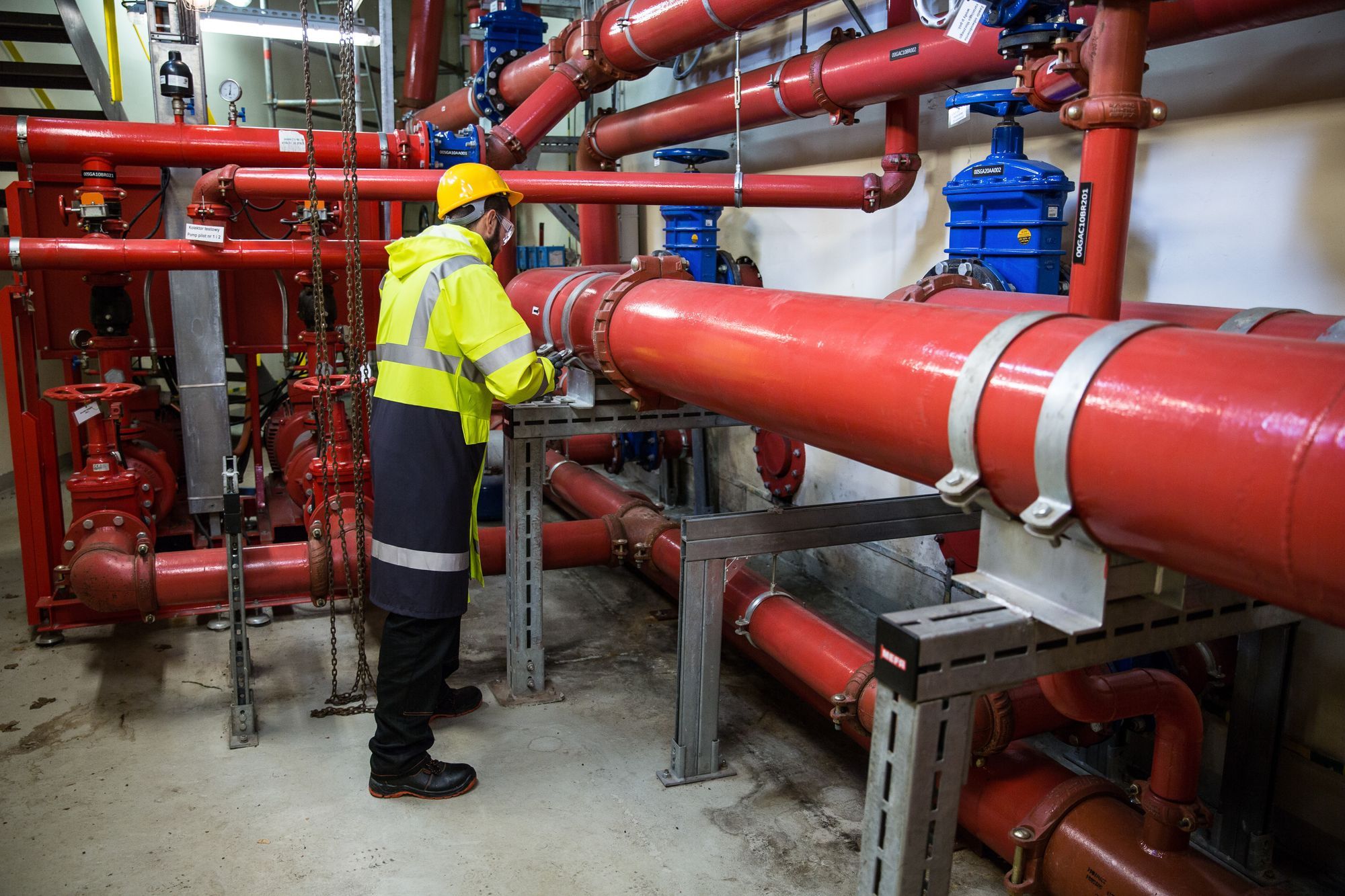 Technician in a hard hat and high-visibility coat inspecting piping and valves in a pump room